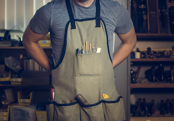 Craftsman in a workshop wearing a gray apron filled with tools such as pencils, a ruler, and a hammer.